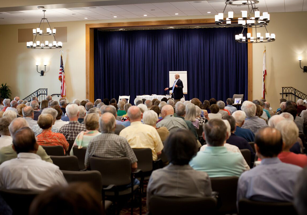 A man standing and giving a presentation in front of several elderly people seated in a large room