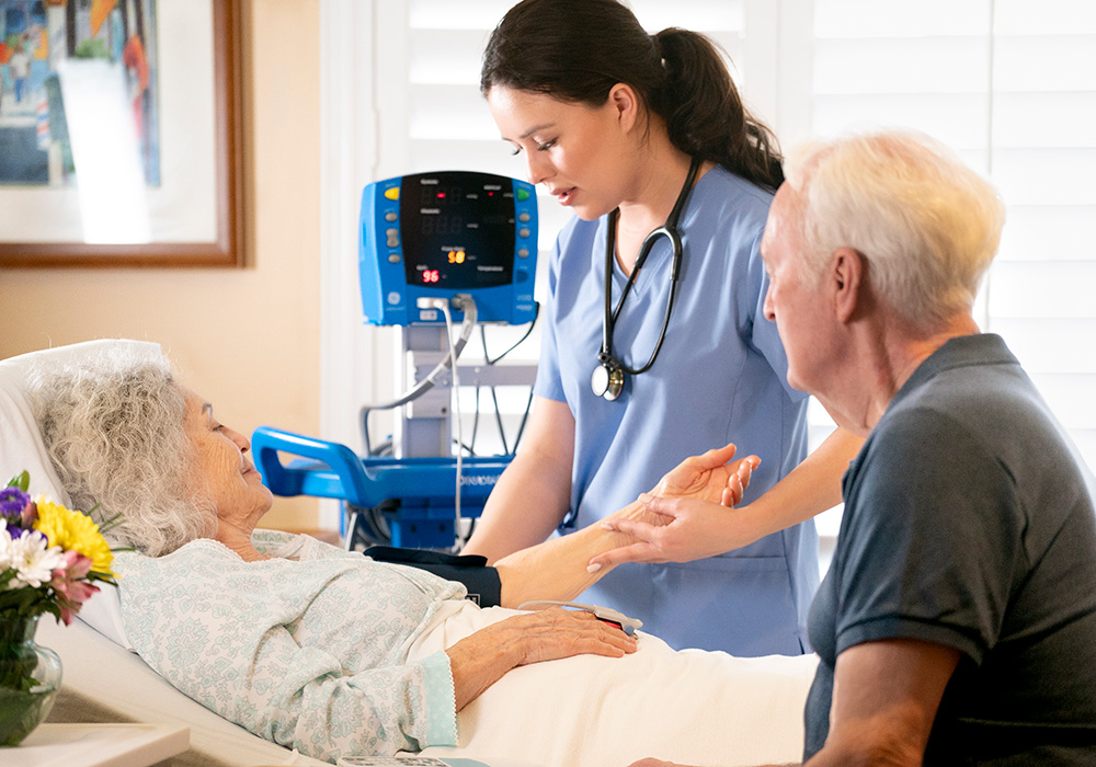 Woman taking blood pressure of another woman laying in bed with a man sitting by her side.