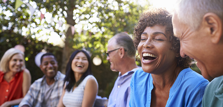 A group of various ages sitting together in a group outside laughing with a large tree above their heads.