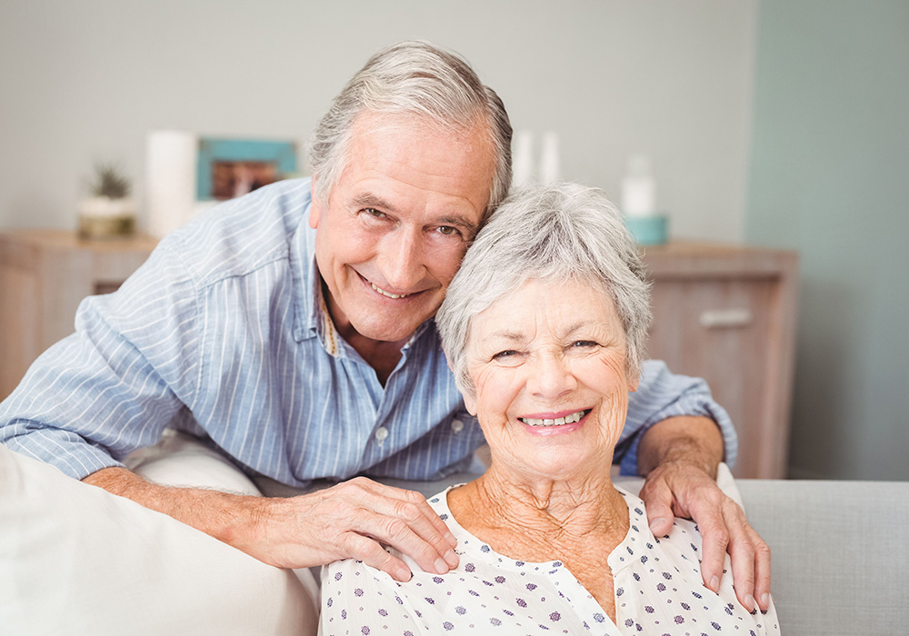 Two seniors smiling together. One senior is sitting on a couch and the other senior is behind her hugging her.