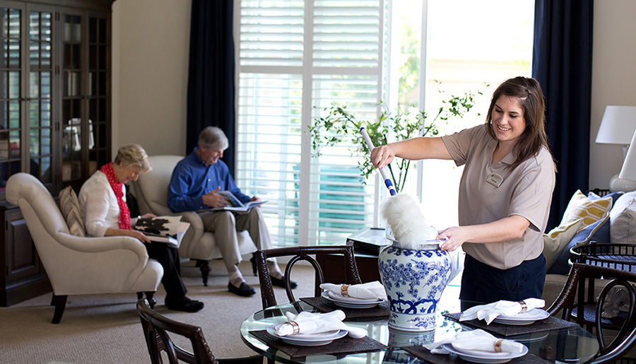 A housekeeper dusting a vase in a home where the seniors who live there are behind her reading a book in chairs.