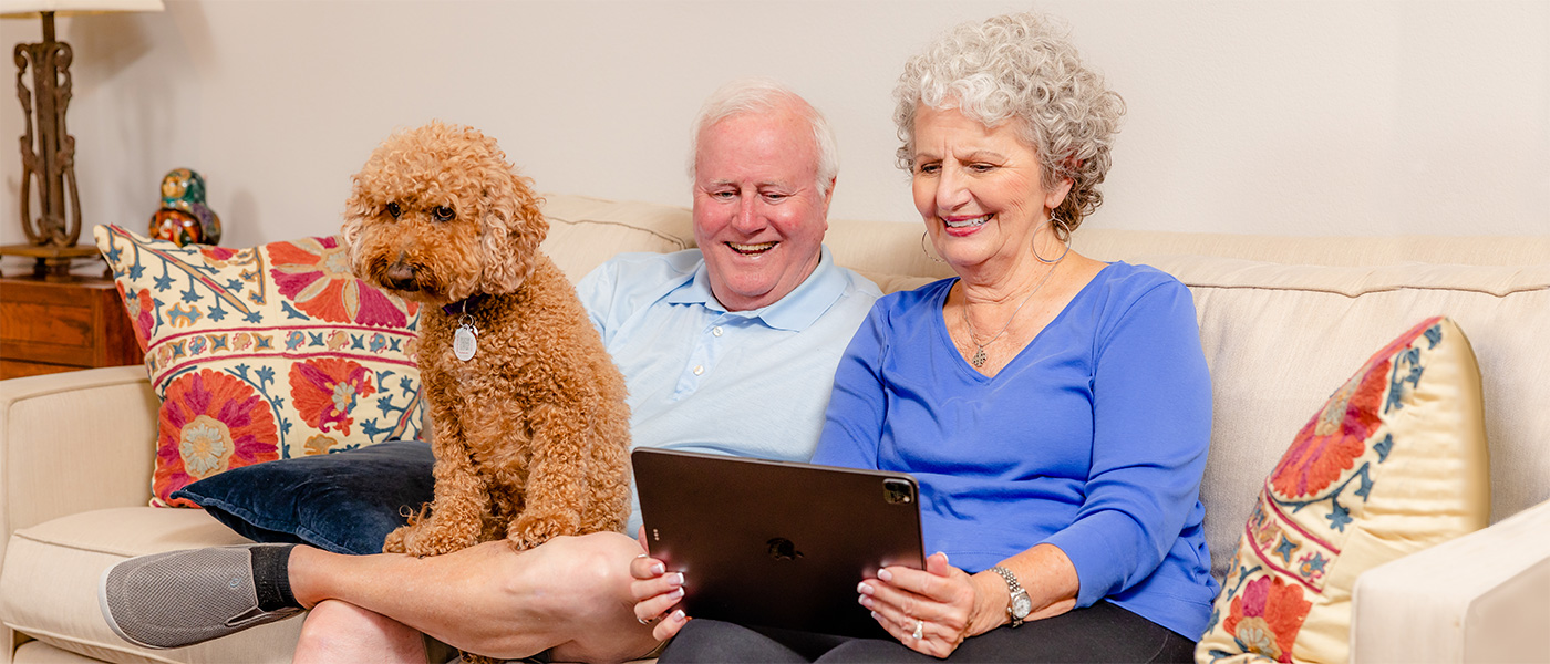 reata-2023-1400x600-35 A resident couple sitting on the sofa looking T laptop with their brown dog