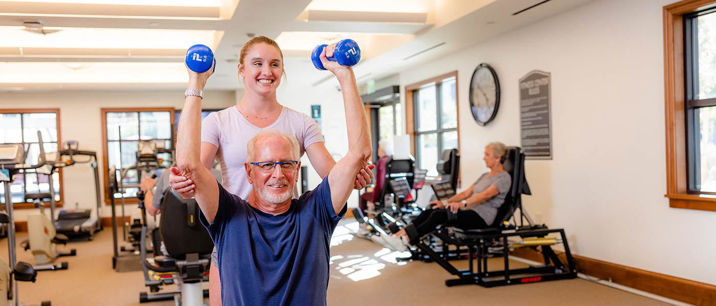 Man lifting weights in a gym being assisted by a woman spotting him with his weights