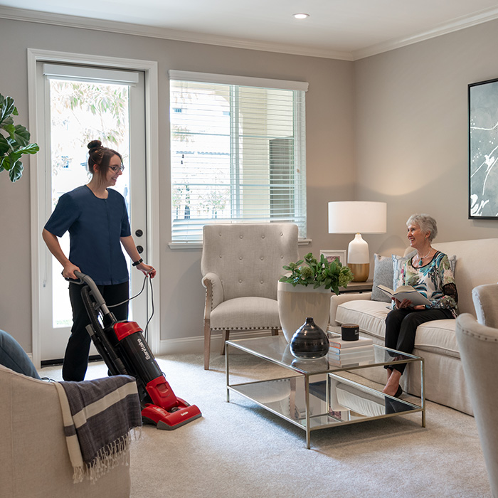 A resident reading a book on the sofa while a woman vacuums the carpet