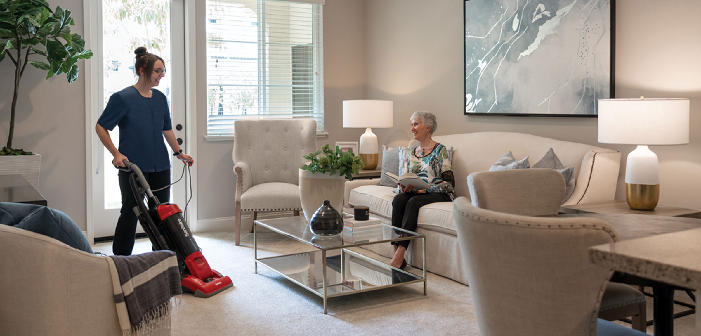 lacosta-2024-600x600-2 A resident reading a book on the sofa while a woman vacuums the carpet