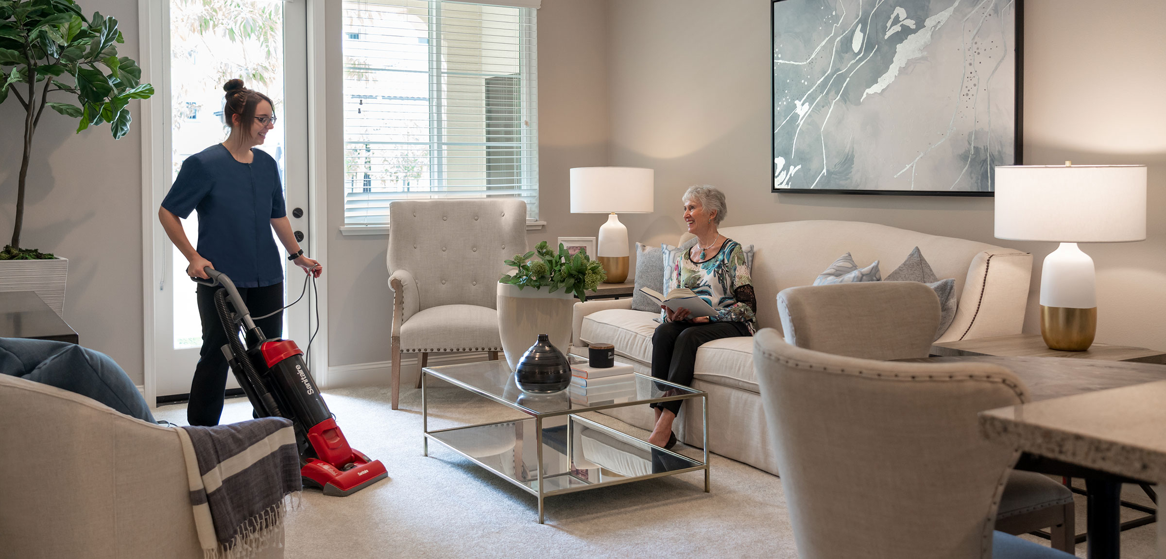 ReataGlen-600x600-13 A woman vacuuming while a resident sits on the couch reading a book